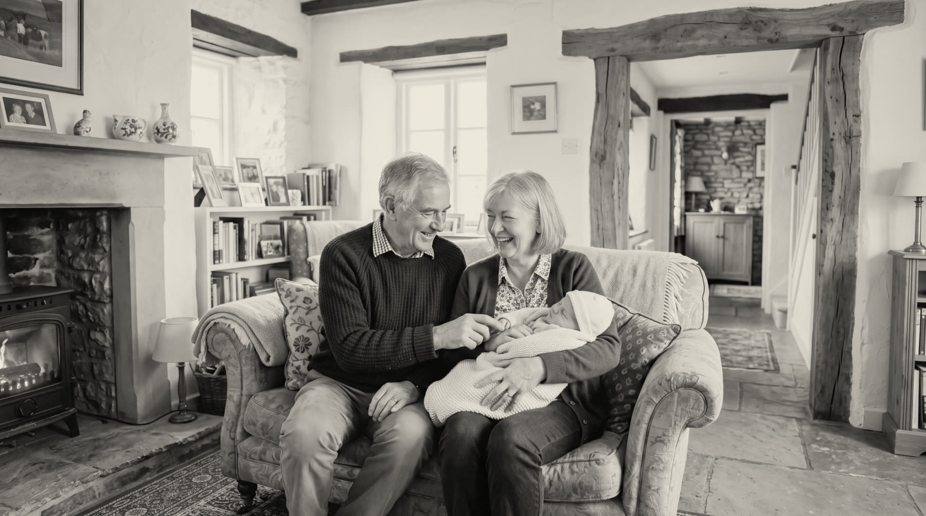 A black-and-white photograph of grandparents sitting together on a sofa in a beamed cottage room, holding a swaddled newborn, a woodburner lit beside them.