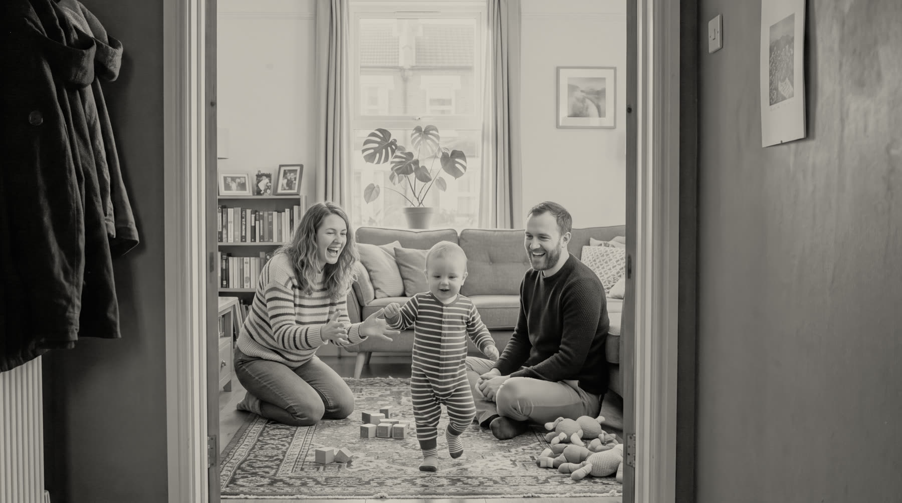 A black-and-white photograph, seen through a doorway, of two parents laughing on the floor of their living room as their toddler takes a few steps between them.