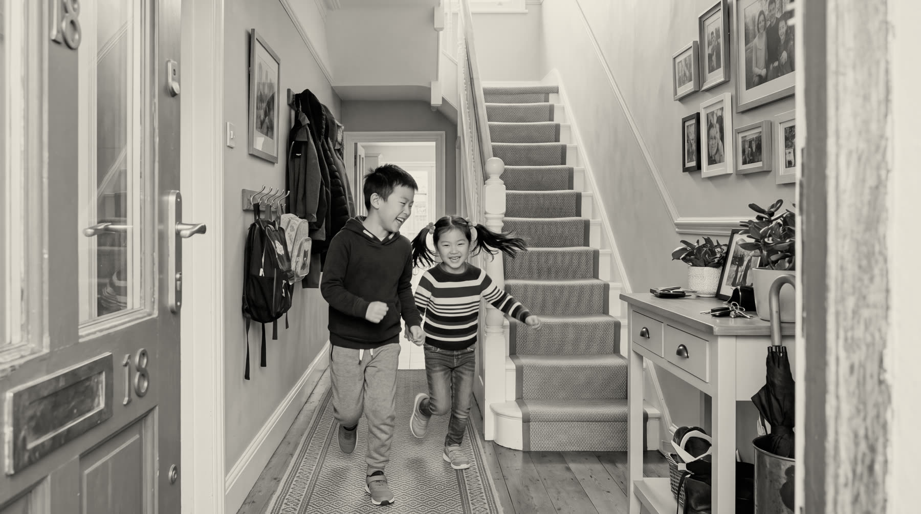 A black-and-white photograph of two children running hand in hand down the hallway of their home, past the front door and the foot of the stairs.